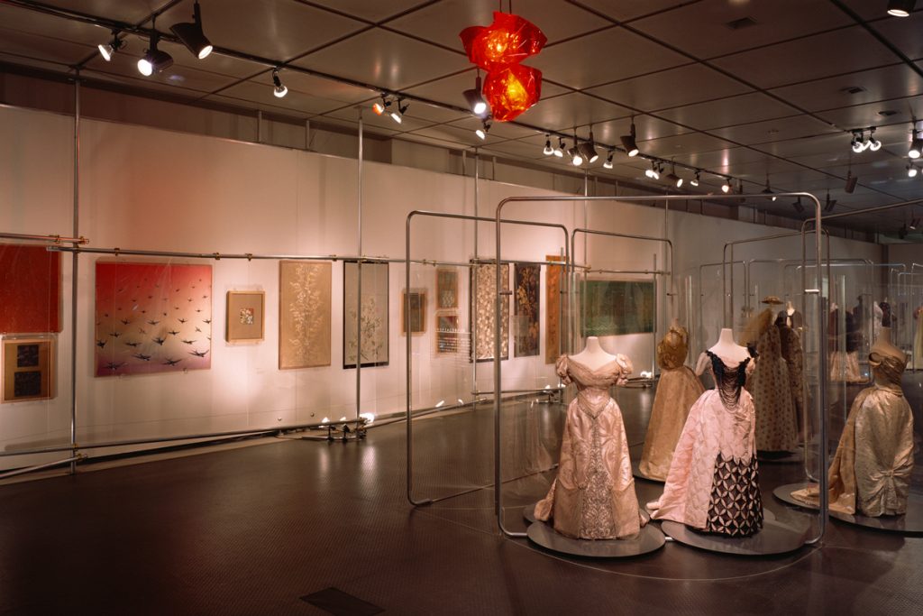 Exhibition display of 19th century feminine gowns and cloth paintings and prints