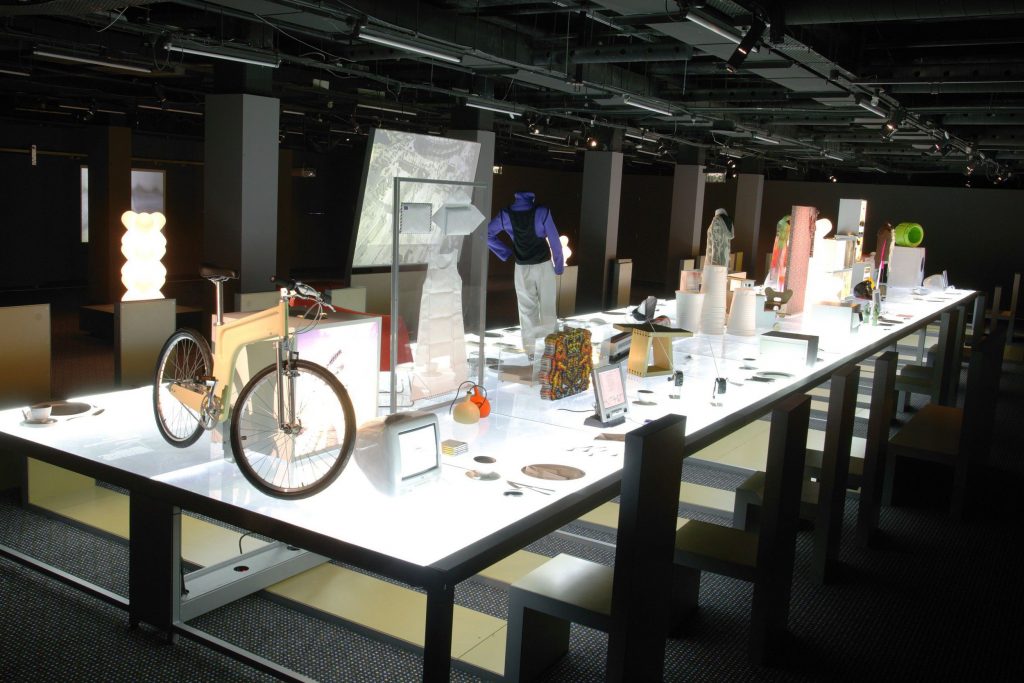 A white-lit table with black frame showcase various exhibits including a bike an Mac computer at the front of the image. The table is surrounded by grey and brown angled chairs.