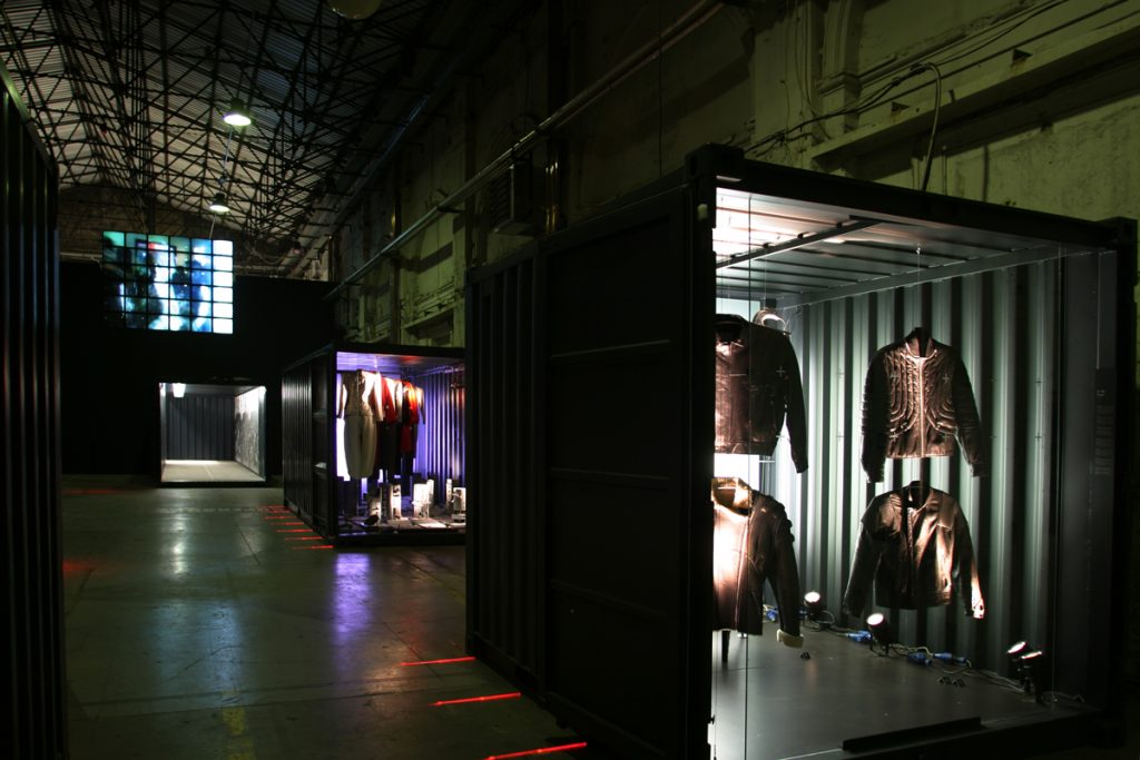 Dimply-lit exhibition display with garments suspended in shipping containers