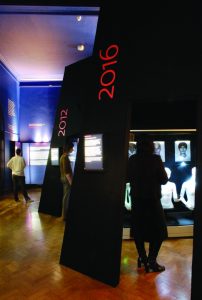 A gallery view showing two sections entitled '2012' and '2016' in large red lettering on black walls. In '2016', are three white mannequins and portrait photos.