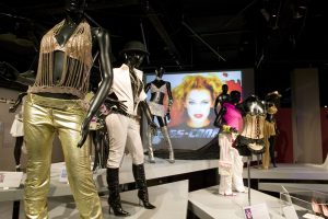 Gallery view showing gold, silver and white outfits worn on black mannequins on white plinths. In the background is a large projector screen with Kylie's face and the word 'SS-CONF' in silver