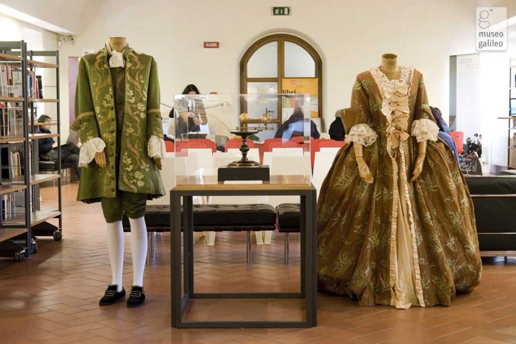 Exhibition display of dressed mannequins in period costume beside scientific apparatus with arched window in background