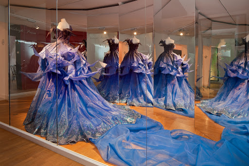 Exhibition display of dressed mannequins in blue dress with a train reflected in mirror in background