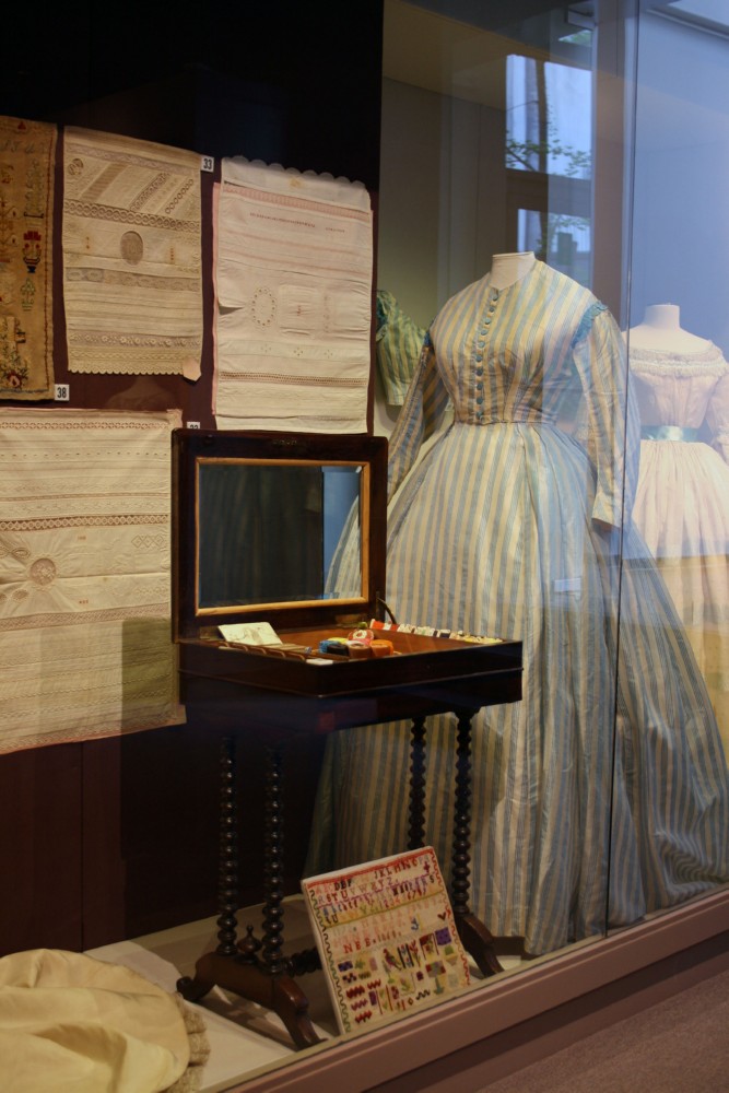 Exhibition display of 18th century gowns with a sewing case in the foreground, and embroidery and samples on the wall and floor