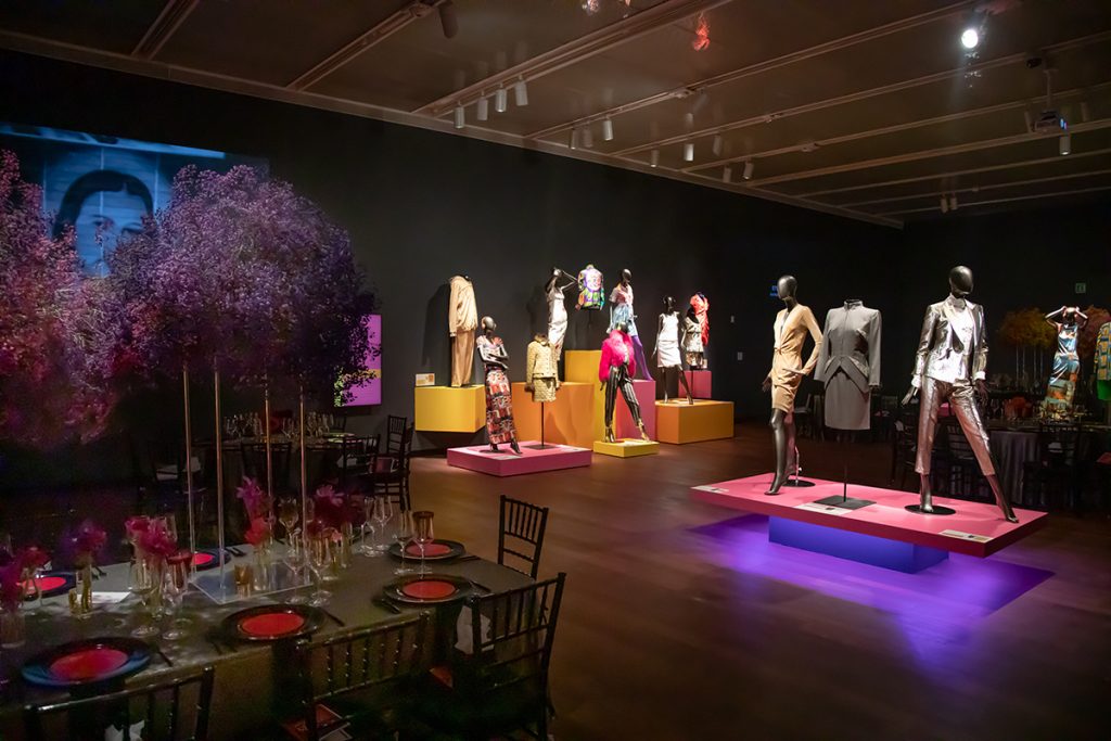 Exhibition display of dressed mannequins on coloured plinths with table and chairs laid for dinner in foreground