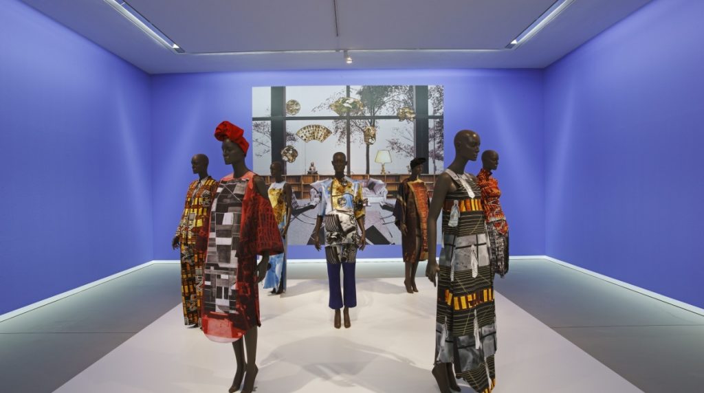 Mannequins wear dresses printed with abstract images of buildings in a blue room. In the background is an illustration of a view from a window.