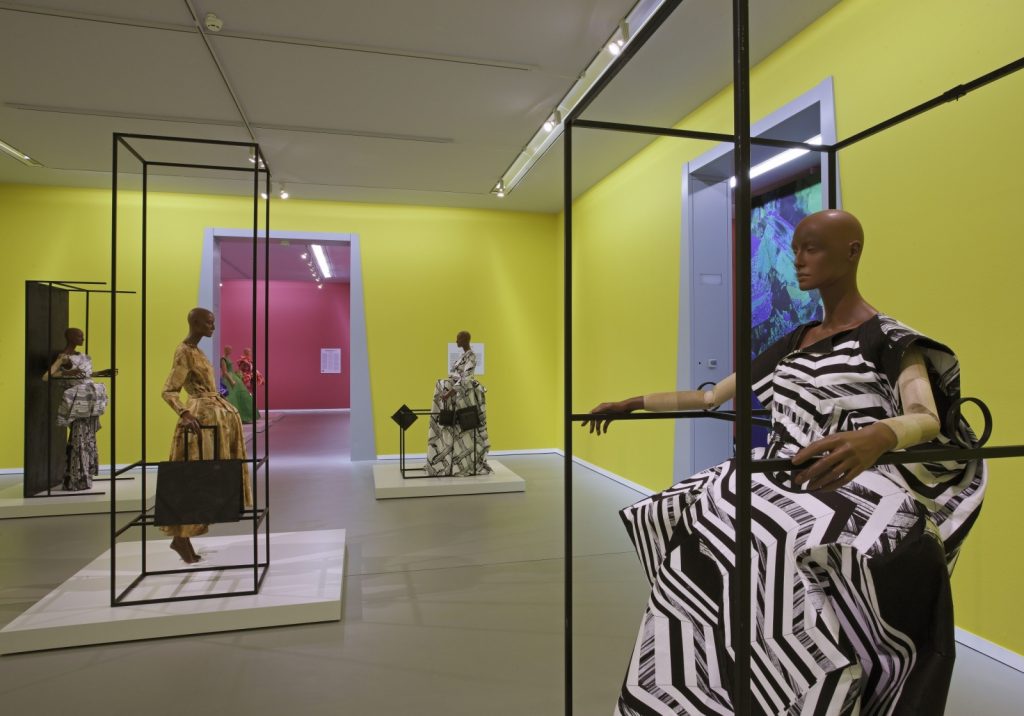 Four mannequins wearing printed dresses stand in or next to black metal framed structuresin a yellow room. In the background is an entry into a pink-painted room.