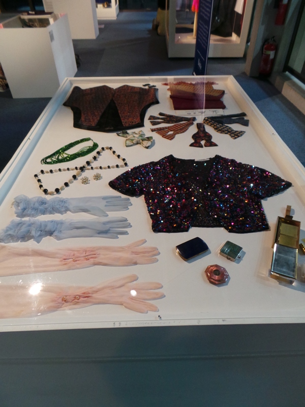 A vitrine displaying clothing pink and blue gloves, a black sequined T-shirt and other accessories.