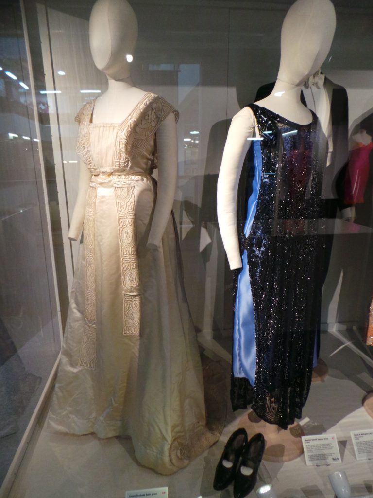 Two female mannequins wearing a pink/ white full length dress and a full length black dress. Black shoes also on display.