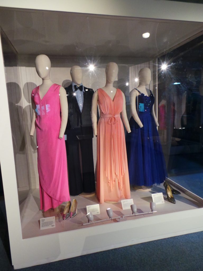 Thre female mannequins wearing evening gowns in hues of bright pink, soft salmon pink and dark blue. Male mannequin wearing a tux.