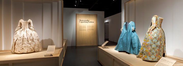 Three dresses incorporating panniers to increase their width, are displayed in front of a panel titled 'Masteworks: Unpacking Fashion'