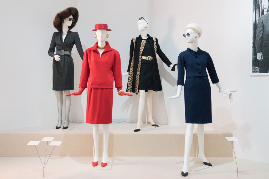 Four mannequins stand with their hands outstretched on two plinths. One wears a grey skirt-suit with fure hat; another a black dress and coat with leopard print trim. In front is a red collared top and skirt with matching boating hat, and a navy skirt with matching jacket.