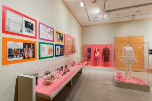 Gallery view showing four dresses and a display of shoes, hats and glasses against a pink backdrop. Above the accessories case is 2D media - posters, illustrations and magazine excerpts, alongside two mini-dresses.