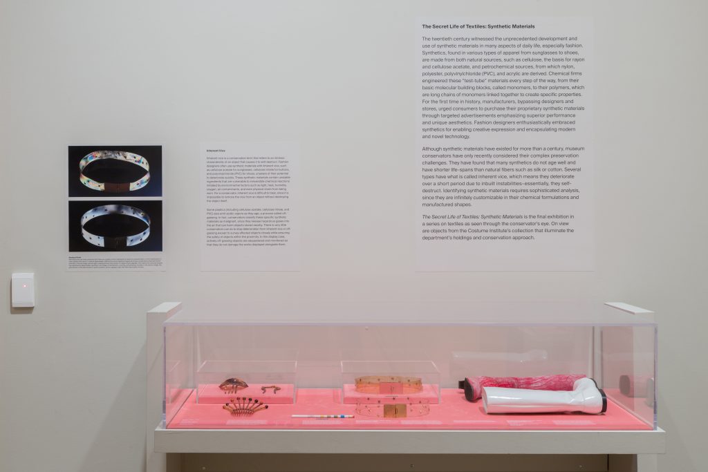 Black exhibition text against a white wall sit above a display case of objects on pink material.