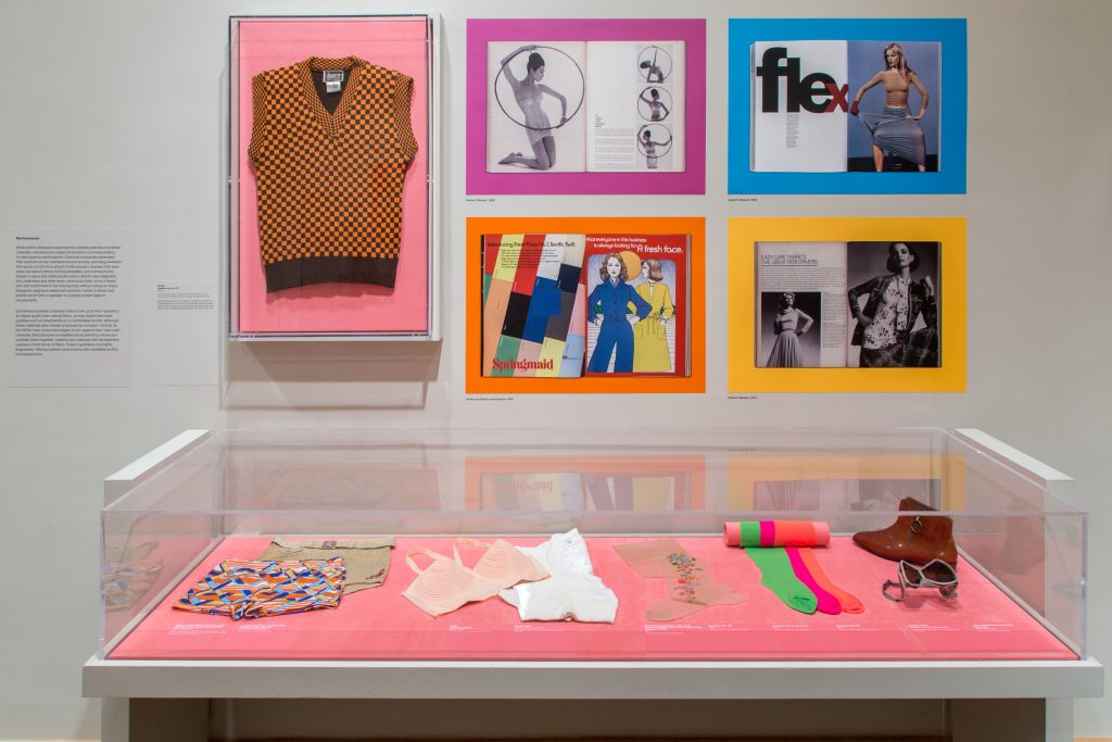 An orange and black checked vest are displayed alongside some 2D graphic media, above a display case of underwear and hosiery.