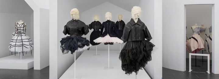 Five black outfits are displayed on tailor's dummies in the foreground, each inspired by black biker jackets and tutus. Each mannequin has a differently styled blonde wig.
