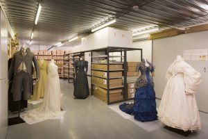 A group of historical European dress, worn on tailors' dummies, stand before archival boxes in a storage space.