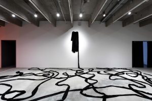 Exhibition display of a garment suspended above a floor with black curvy lines on the floor