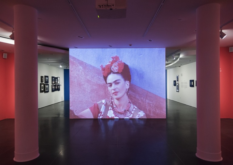 A photograph of Frida Kahlo on a screen and has decorative flowers in her hair. Outfit is complete with red and white necklaces and a red top.