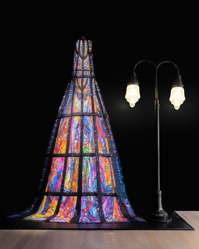 Exhibition displaying a long Michael Cinco dress next to a lit lamppost.