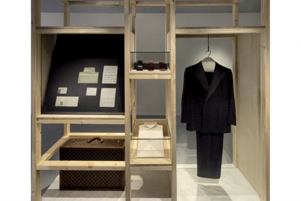 Exhibition display of shelving with garments and belongings hanging and mounted on shelves