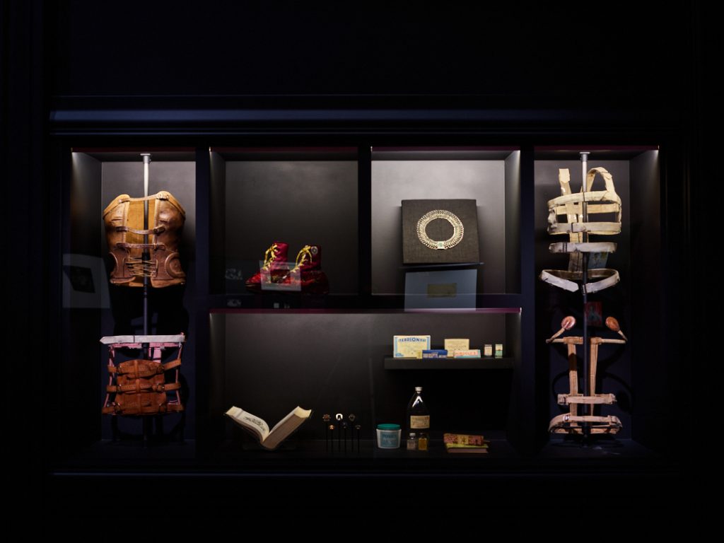 A display of objects belonging to Frida Kahlo such as a silver collar necklace, open book, plaster corsets, braces, red boots with yellow laces, and various sizes of brown, white and yellow bottles and packages.