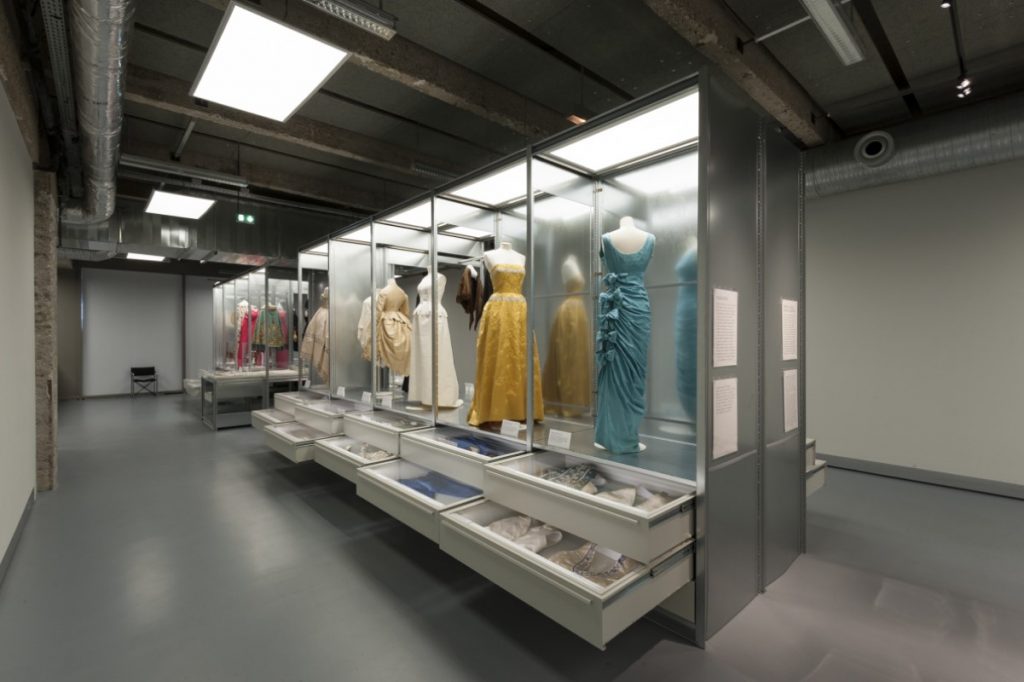 Exhibition display of dressed mannequins in case with open drawers beneath
