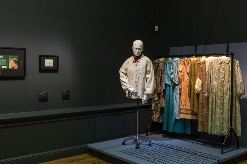 Exhibition display of dressed mannequin with 3d printed head in front of a rail of garments