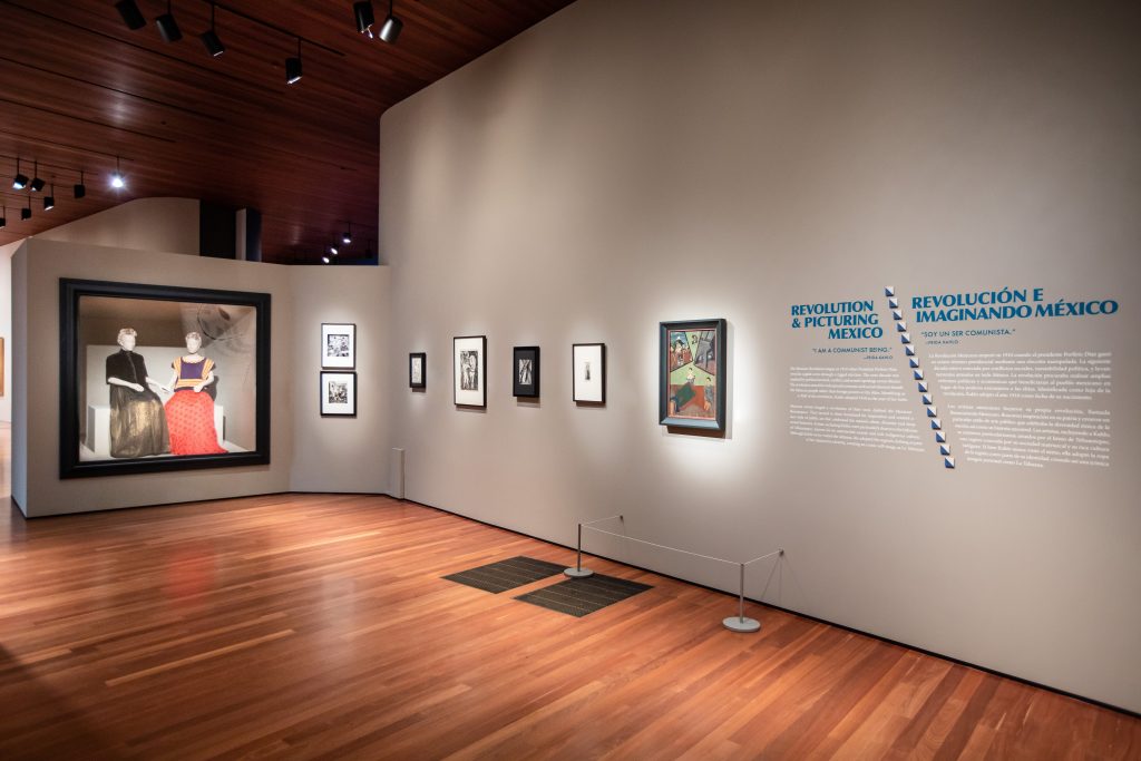 A display of painting and photographs. A vitrine displaying two female mannequins fixed in a seated position. (Left) wears a black top paired with a long silver skirt. (Right) wears a black sleeveless top with an orange/ peach long skirt.