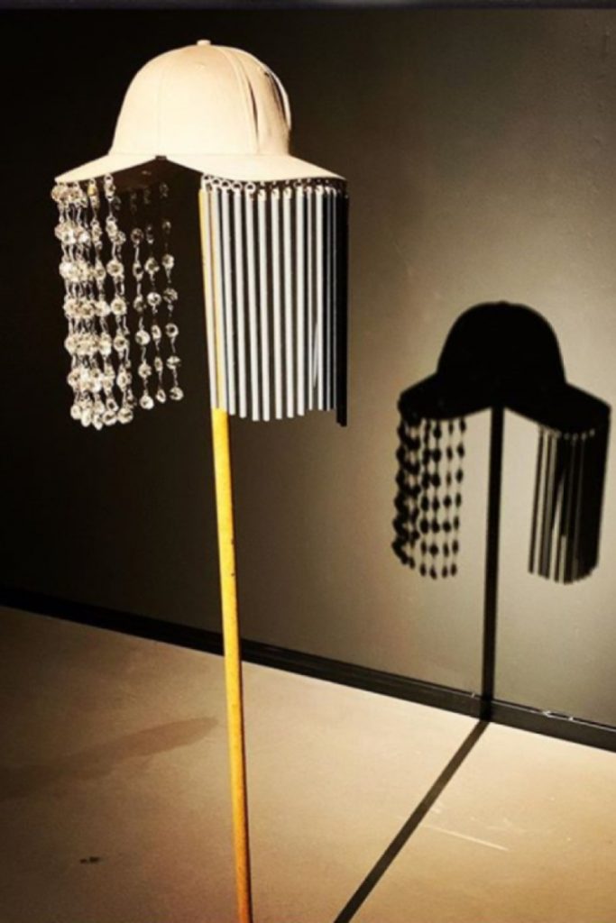 Exhibition display of hat with wind chime fringes and backed by silhouette