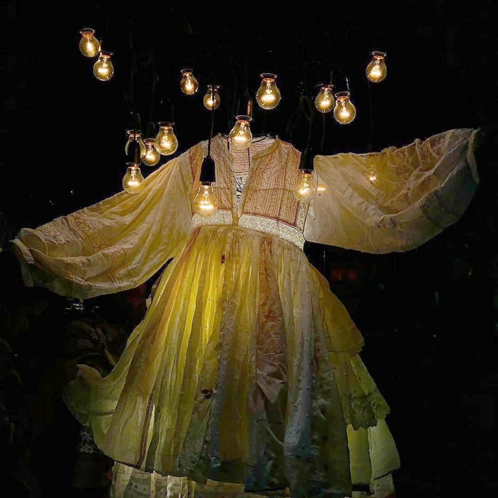 Exhibition display of white diaphanous dress lit up with lightbulbs overhead
