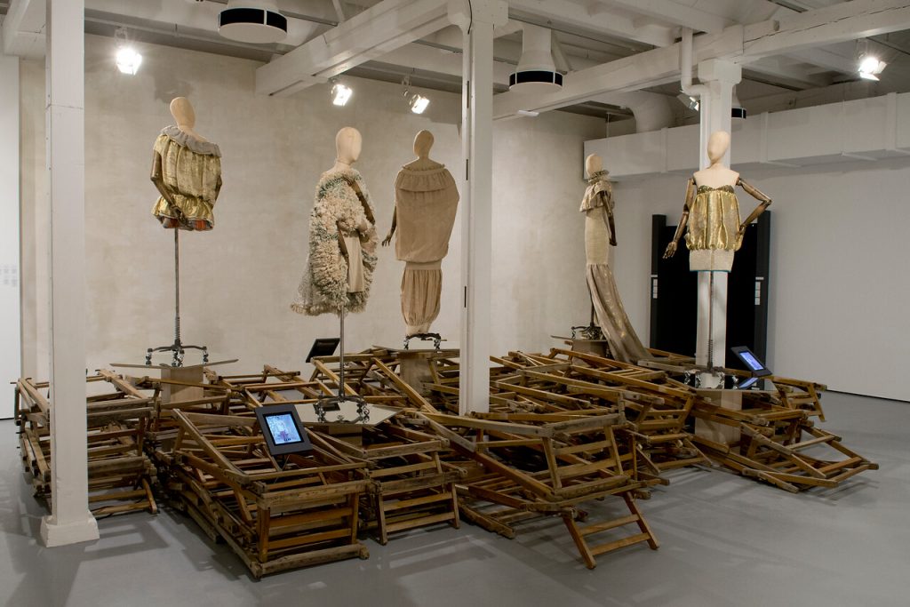 Exhibition display of dressed mannequins above folded wooden deckchair frames