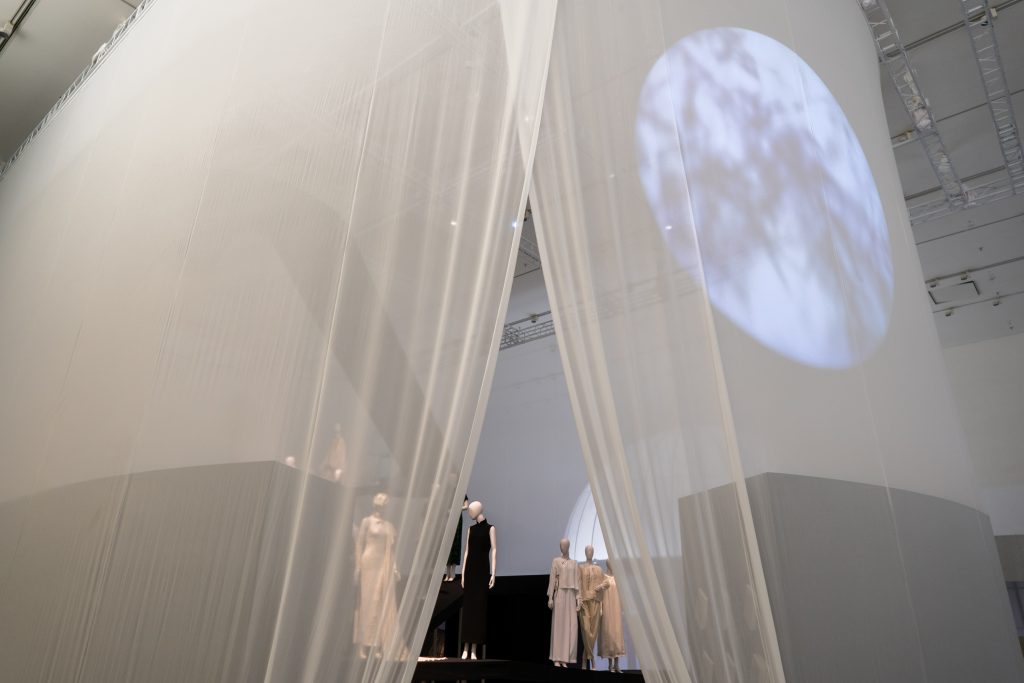 Exhibition display of dressed mannequins with projection of the moon and looking through gap in net curtain