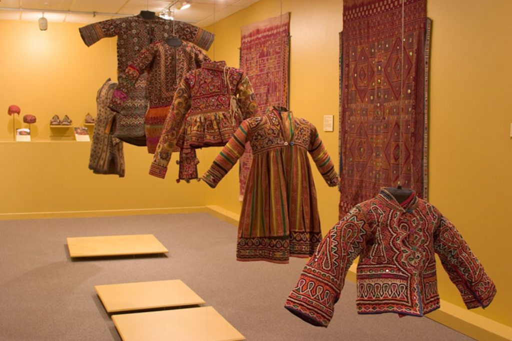 exhibition display of dress suspended from ceiling , textile panels hung against yellow painted walls