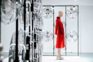 A red PVC coat worn on a crescent-patterned mannequin, set against a display wall of silver air fans