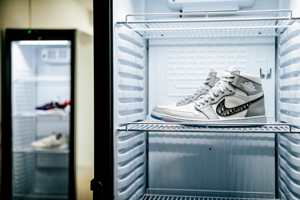 A pair of Dior x Nike high-top trainers sit on a fridge shelf. In the background is another fridge with two more pairs of trainers.