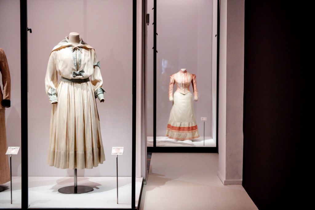 Two 18th century cream dresses with pale green and pink detailing, set in black-framed display cabinets and pink walls.