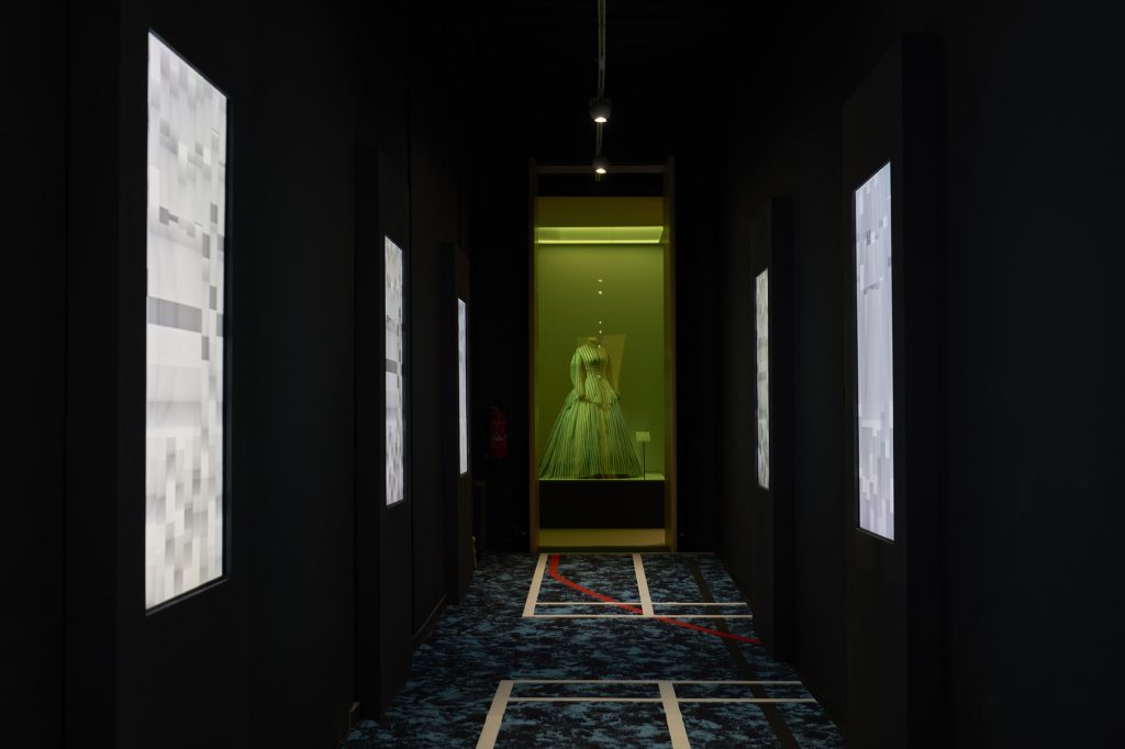 An 18th dress is framed in a green-glassed cabinet, surrounded by screens. The floor is an abstracted basketball court.