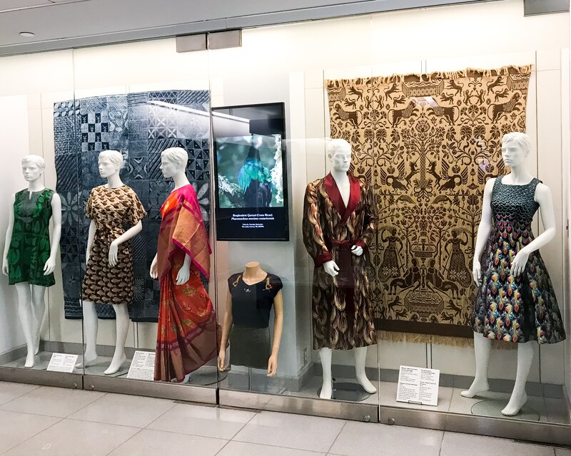 Exhibition display of dressed mannequins in glass case with pattern fabric in background