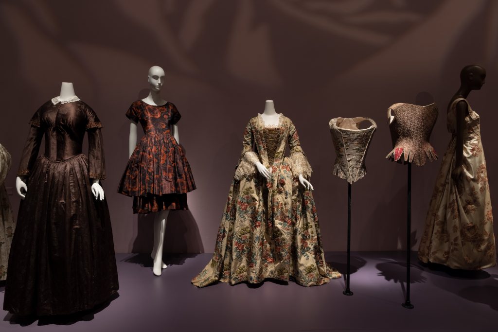 Exhibition display of dressed mannequins in historical rose-patterned garments including corsets