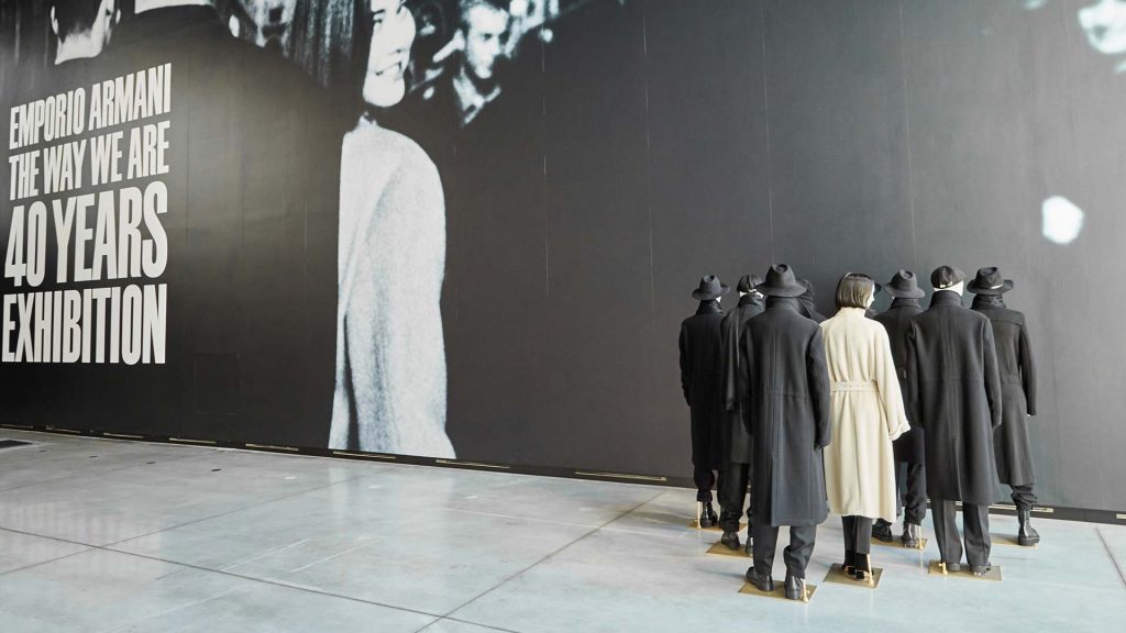 A group of mannequins stand in a triangular formation facing a wall and wearing dark suit jackets and hats. In the centre is a female mannequin with a cream trench coat.