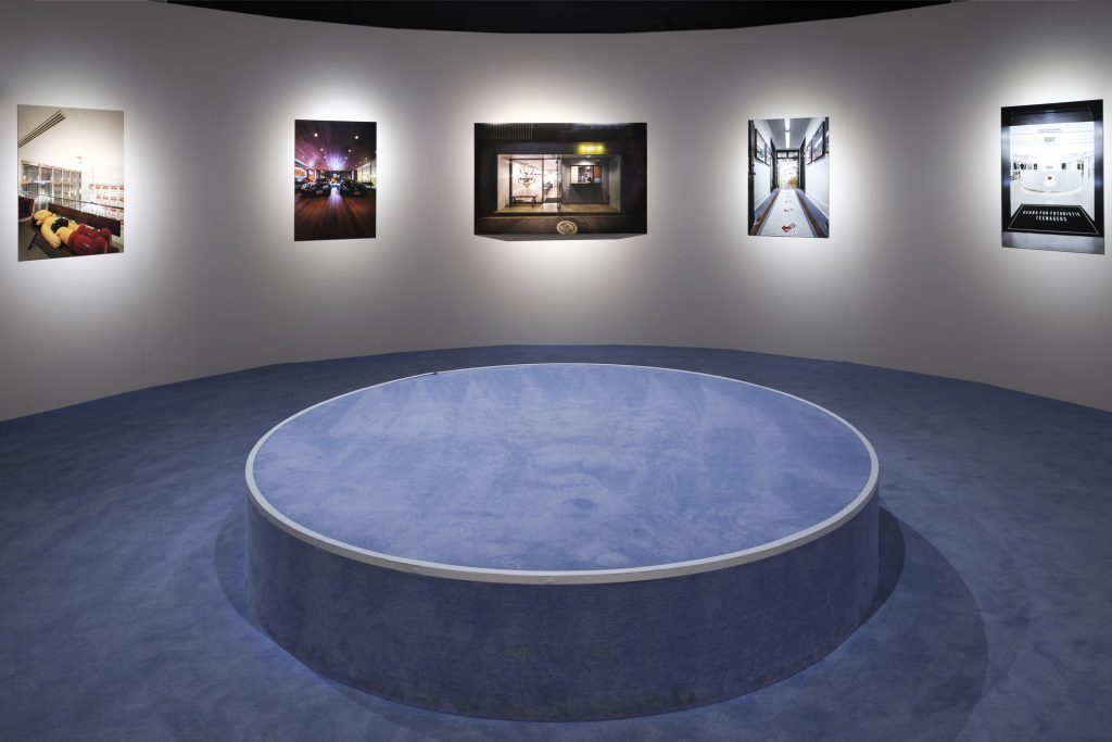 Exhibition display of pictures in a round space with round podium in foreground covered with blue carpet