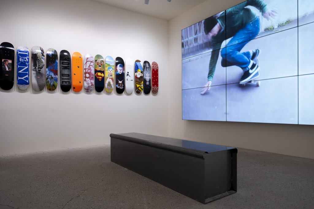 Exhibition display of skateboards on the wall and large screen showing skateboarding film