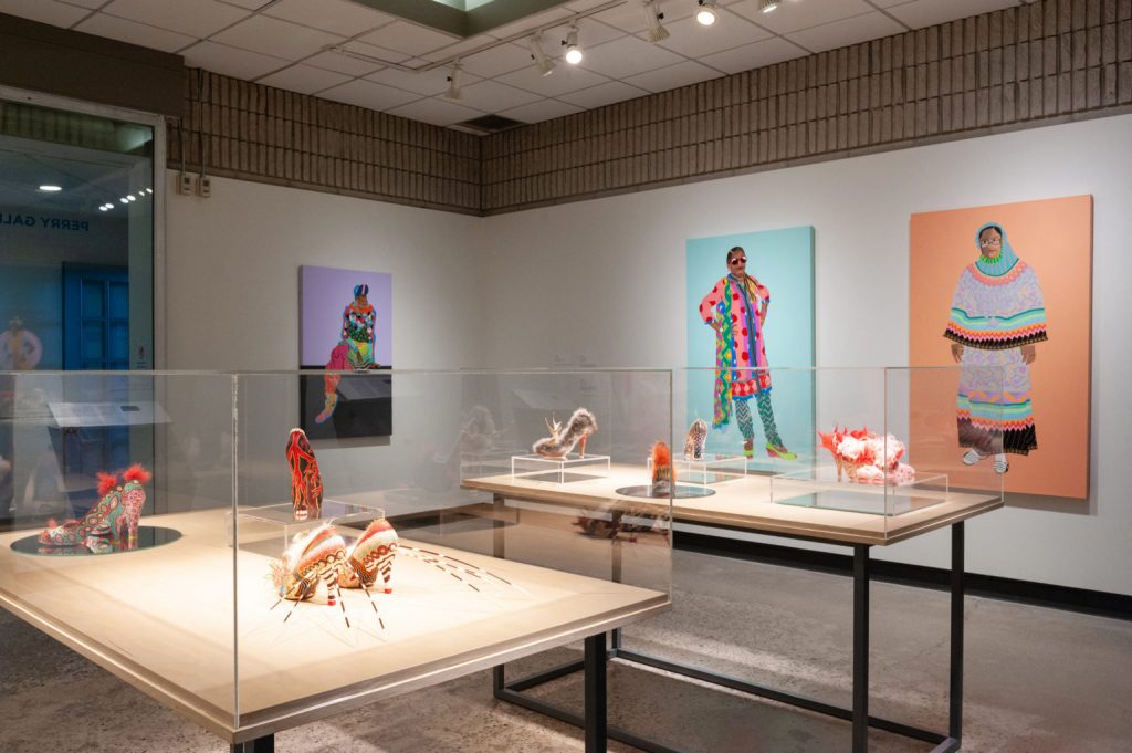 Exhibition with two standing glass cabinets displaying footwear, and three paintings; one on one wall, two on the next wall.