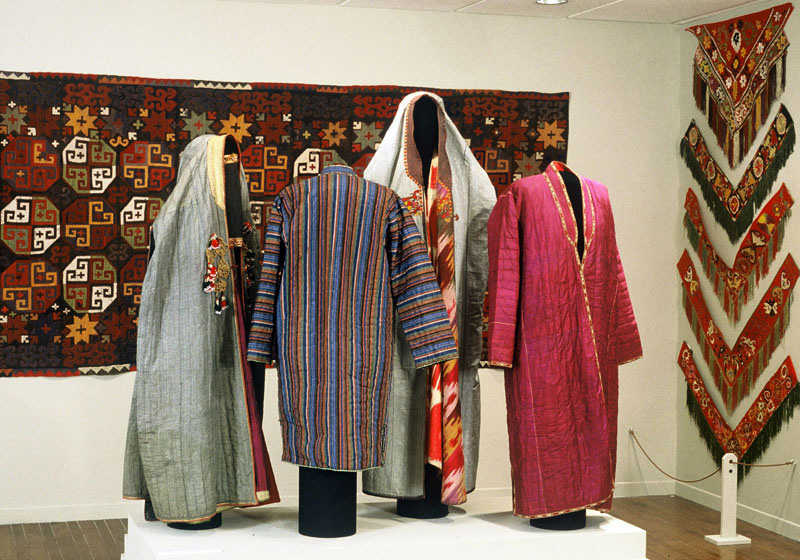 Exhibition with mannequins on a plinth displaying garments from Afghanistan with textiles hanging from the wall.