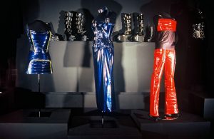 Exhibition with three mannequins displaying garments, and three shoes.