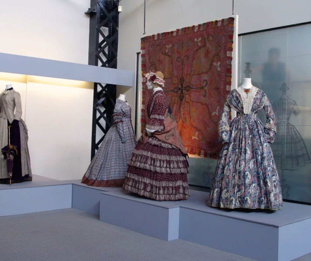 Exhibition with four mannequins displaying period dresses with printed, embroidered or woven paisley.