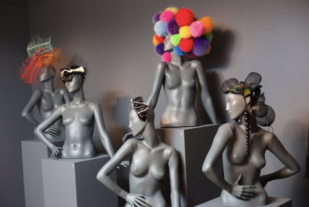 Exhibition with the torsos and heads of mannequins on plinths wearing hats.