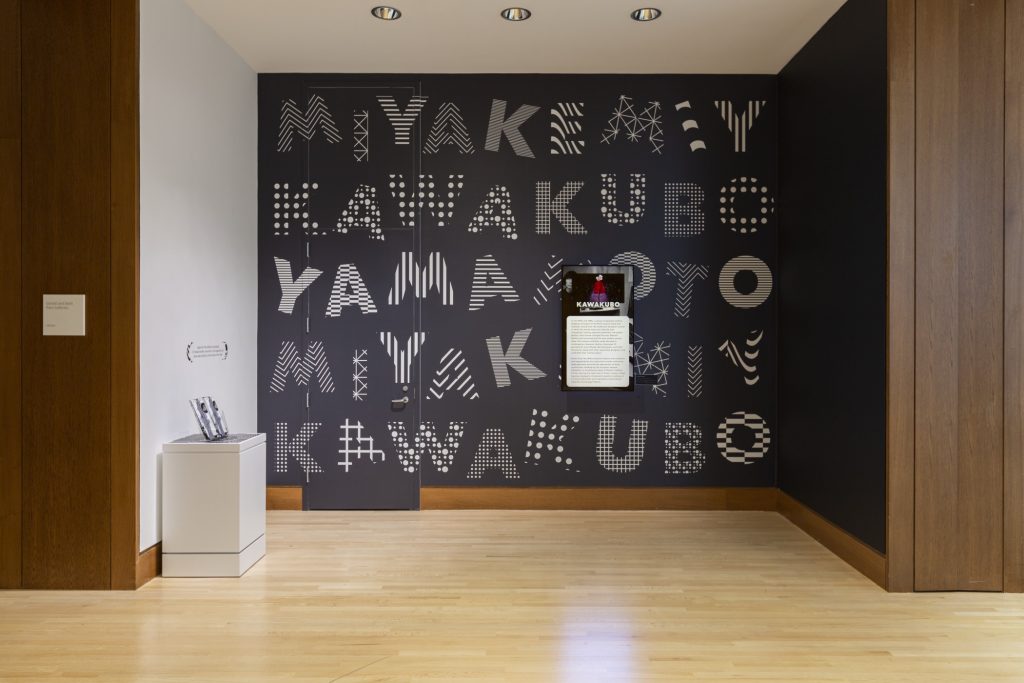 Exhibition with black wall with graphic white letters spelling Miyake, Kawakubo and Yamamoto, a text panel and a plinth with leaflets.