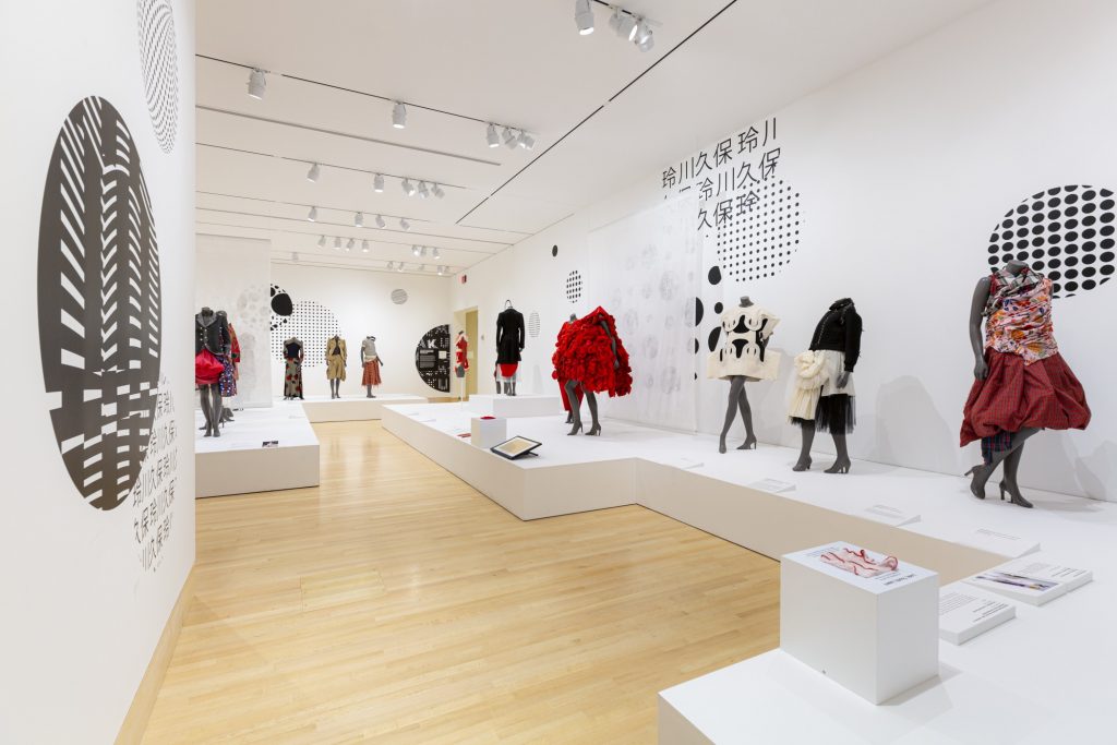 Exhibition with white walls and graphic decorations with plinths holding mannequins displaying garments.